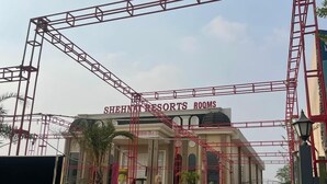 Entrada do Shehnai Resorts, com um portão de metal vermelho e dois carros estacionados na frente.