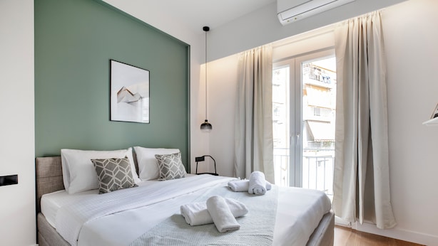 Un letto rifatto con cura, con copripiumino bianco e due cuscini, un quadro appeso al muro e una finestra con tende.