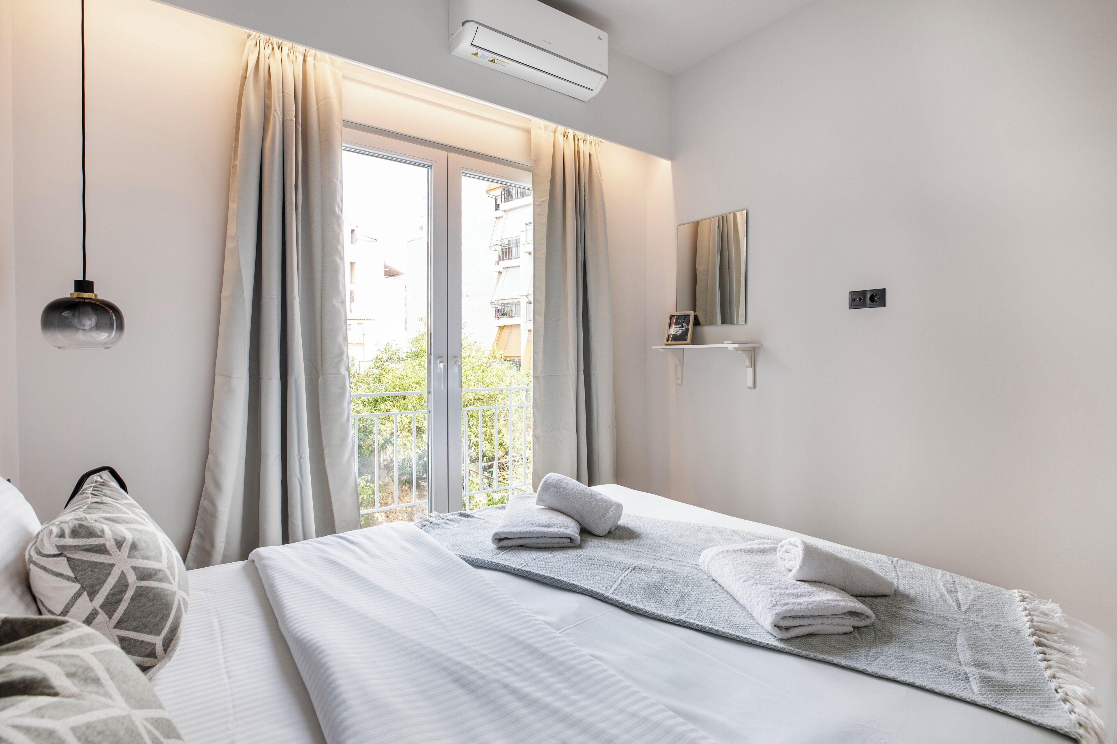 A hotel room with a bed, a window with curtains, a wall-mounted air conditioner, a bedside table with a lamp, and a view of buildings outside.