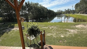 A wooden deck with a metal railing overlooking a calm lake, surrounded by trees and a grassy area.