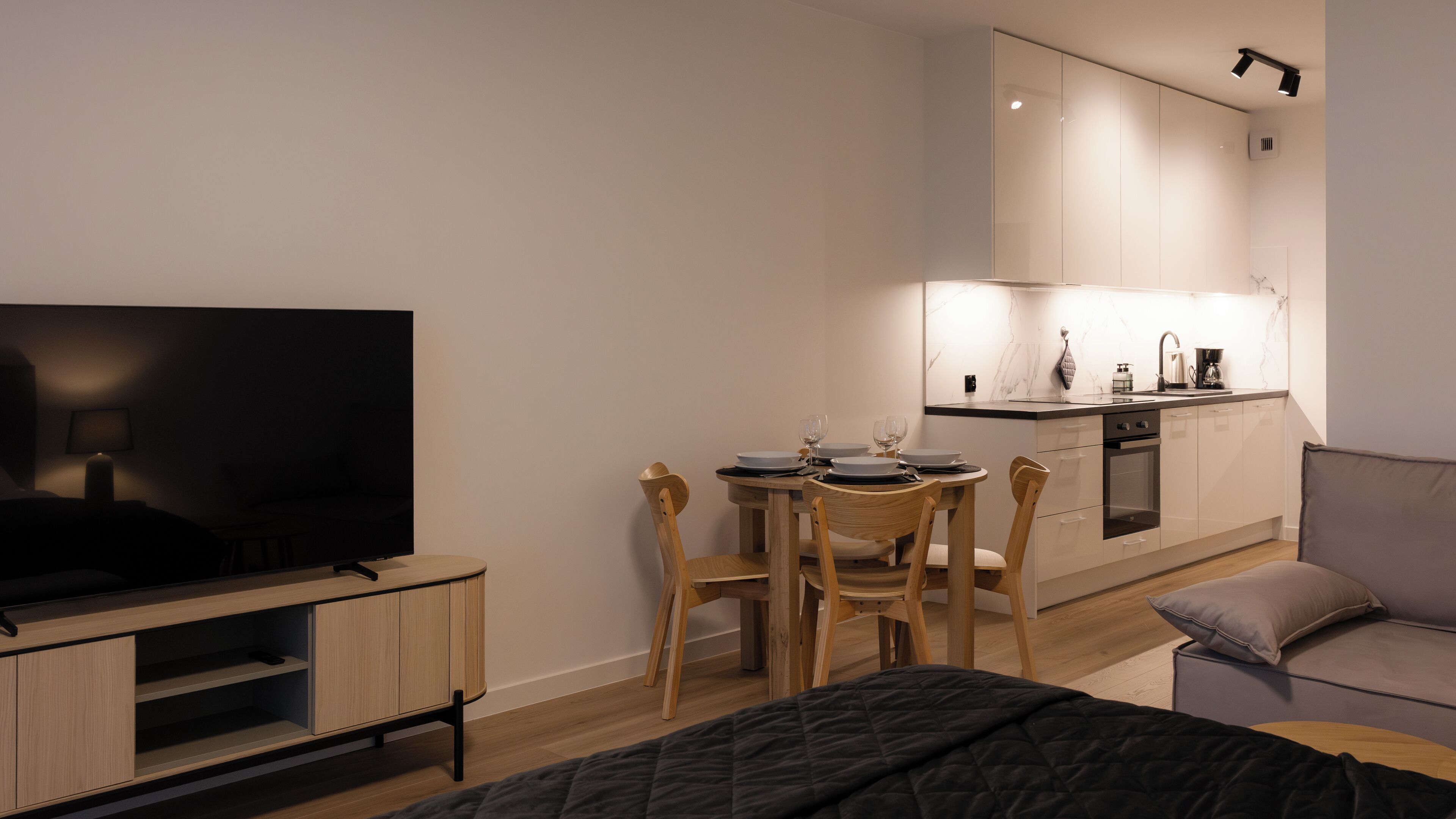 A modern hotel room with a TV, a dining table with chairs, a sofa, and a kitchen area with a sink and stove.