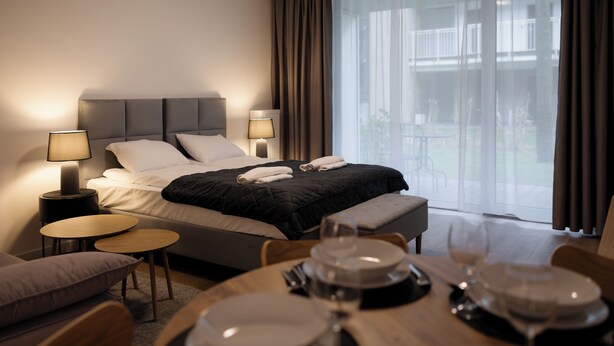 A modern hotel room with a bed, bedside tables, a round dining table with plates and glasses, and a view of the outdoors. - Apartamenty Sun & Snow Nadmorska Florina (Leba)