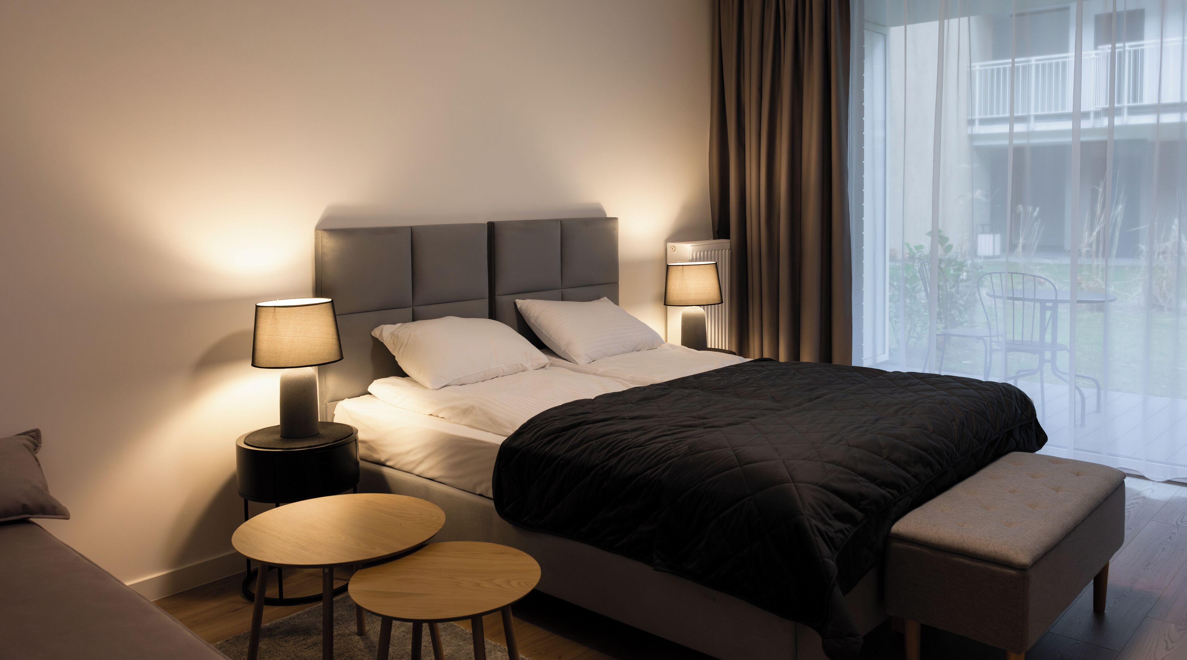 A modern hotel room with a large bed, two bedside tables, a small round table, a bench, and a view of the outdoors.