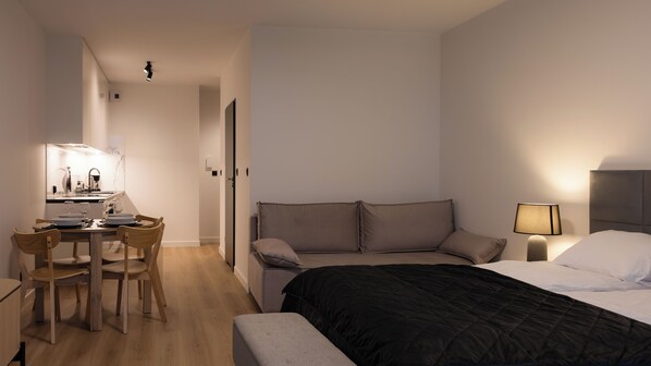 A modern hotel room with a bed, bedside table, lamp, and a small desk with a chair and a mirror.