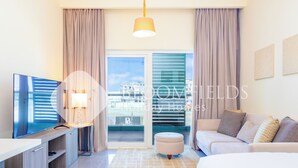 A modern hotel room with a flat-screen TV, a sofa, an ottoman, and a large window with curtains. - Bloomfields Cozy STU Balcony Leonardo (Abu Dhabi)