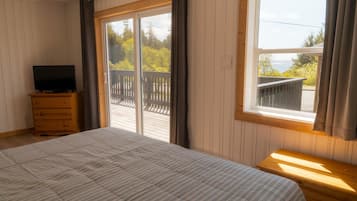 A bedroom with a bed, a dresser, a TV, and a large window with a view of a deck and trees.