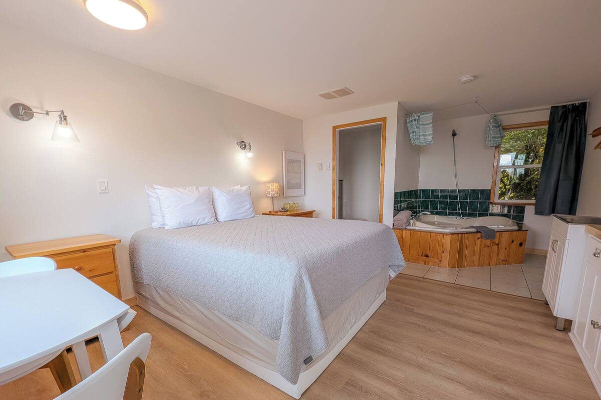 Une chambre à coucher avec un lit, une table de chevet et un petit bureau avec une chaise. Il y a une salle de bain avec un lavabo, un miroir et une serviette accrochée au mur.