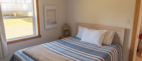 A bedroom with a bed, a window, a framed quote on the wall, and a nightstand with a lamp.