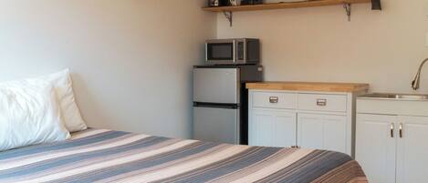 Un dormitorio con cama, microondas, refrigerador y una pequeña kitchenette con pileta y alacenas.