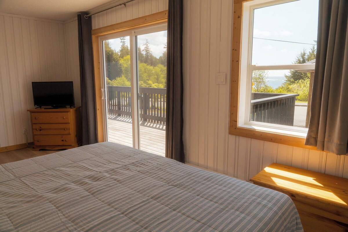 A bedroom with a bed, a television on a wooden dresser, and a large window with a view of a deck and trees.