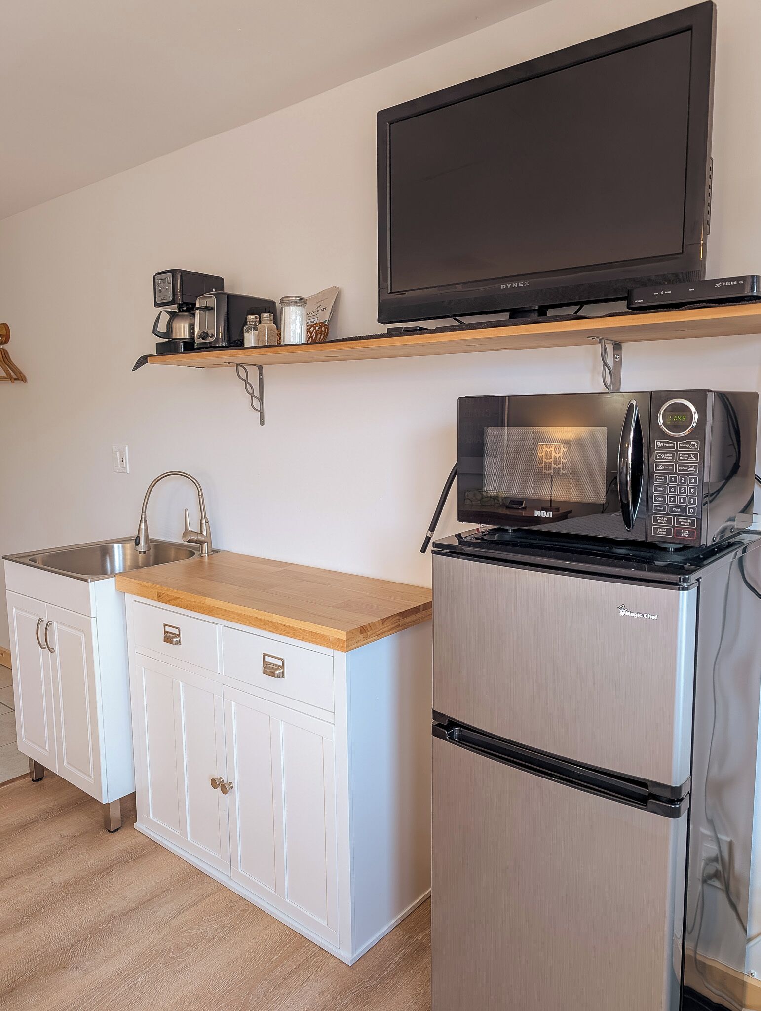 A compact kitchen with a microwave, a refrigerator, a sink, and a television mounted on a shelf.