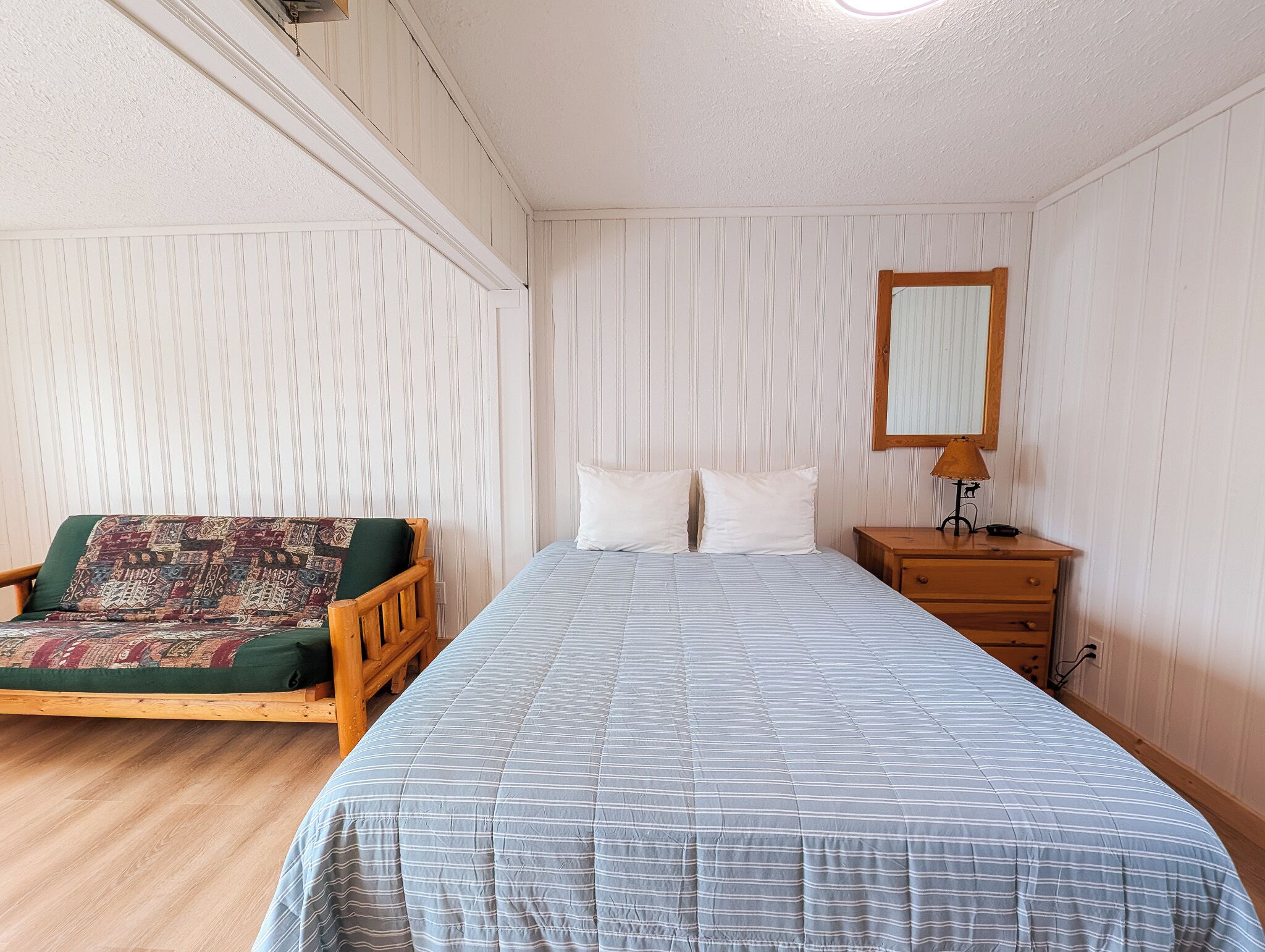 A room with a bed, a wooden chair with a patterned cushion, a bedside table with a lamp, and a mirror on the wall.