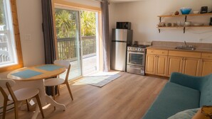 A compact kitchen with a refrigerator, stove, and wooden cabinets. There is a dining area with a table and chairs, and a balcony with a view of palm trees. - Little Beach Resort (Ucluelet)