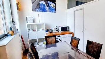 Een eethoek met een houten tafel en stoelen, een magnetron, een klok en een plank met keukenbenodigdheden.