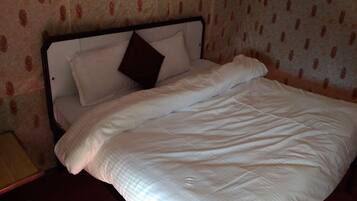 A bed with white bedding and a brown pillow, a wooden nightstand, and a pair of green slippers on the floor.