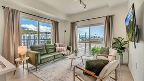 Un salón espacioso con un sofá verde, una mesa de centro blanca y vistas a un paisaje urbano a través de amplias ventanas.
