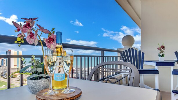A balcony with a table set for a drink, featuring a bottle of Magdolina, a glass, and a decorative orchid.