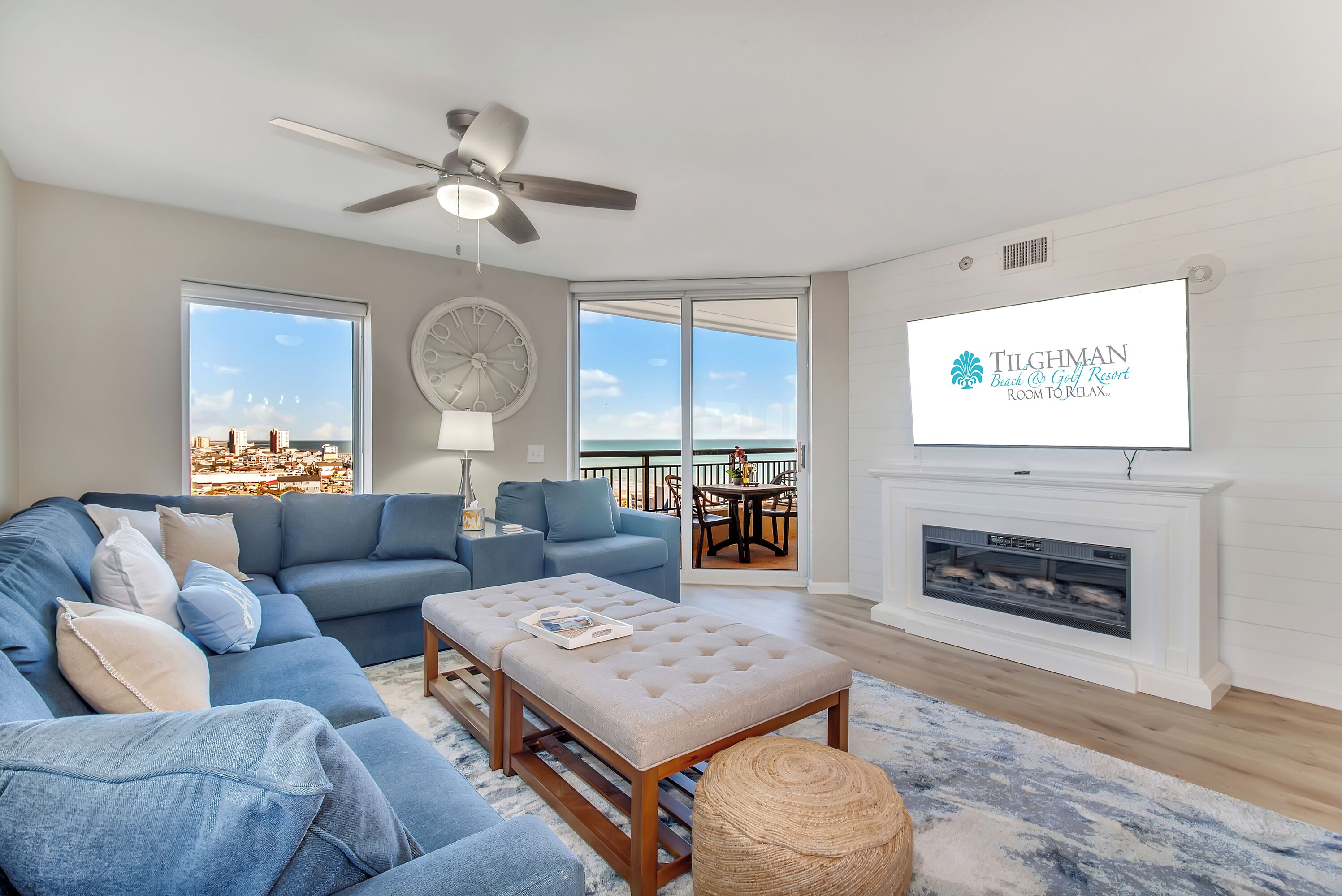 A living room with a blue sofa, a beige ottoman, a ceiling fan, a fireplace, and a TV displaying the Tilghman Beach & Golf Resort logo.
