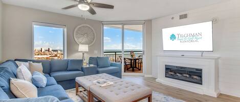 A living room with a blue sofa, a beige ottoman, a ceiling fan, a fireplace, and a TV displaying the Tilghman Beach & Golf Resort logo.