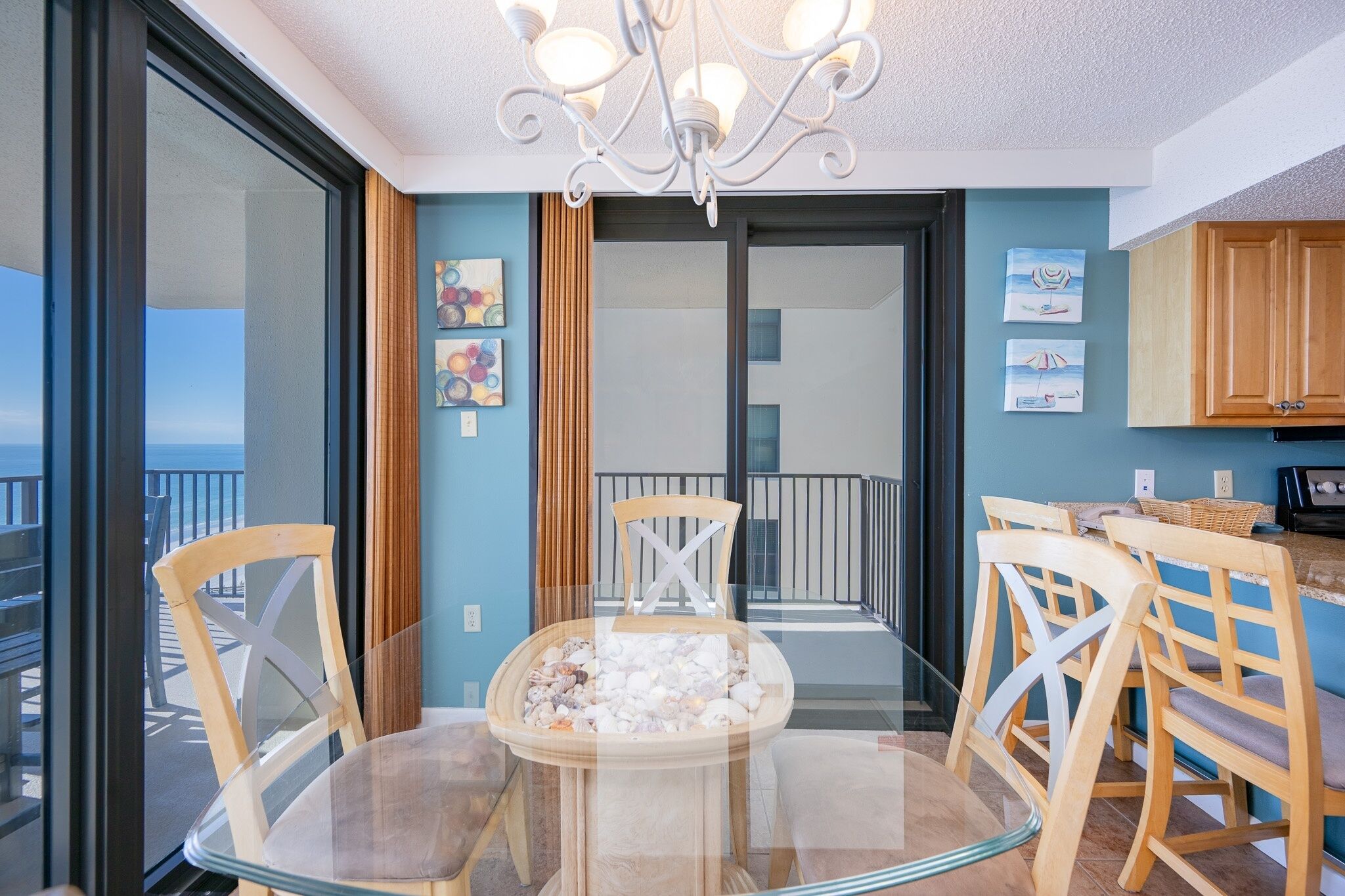 A balcony with a glass table and chairs, a view of the sea, and a kitchen area with wooden cabinets.