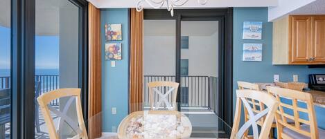 A balcony with a glass table and chairs, a view of the sea, and a kitchen area with wooden cabinets.