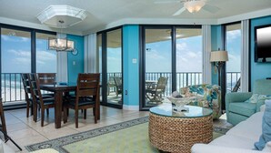 A spacious living area with a dining table, chairs, a round coffee table, and a sofa. There is a large window offering a view of the ocean. - Phoenix Iii 3059 3 Bedroom Condo (Orange Beach)