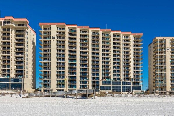 A high-rise hotel with multiple balconies, a sandy beach in the foreground, and a clear blue sky. - Phoenix Iii 3059 3 Bedroom Condo (Orange Beach)