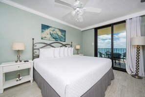 Une chambre d’hôtel avec un lit, des tables de chevet, un ventilateur de plafond, un tableau et un balcon avec des chaises.