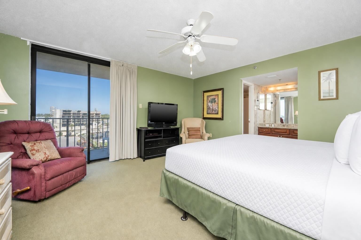 Una habitación de hotel con cama, televisión, sillón, ventilador de techo, balcón con vistas y baño con espejo.