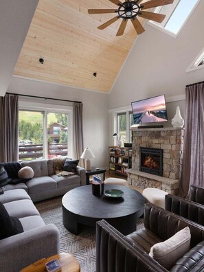Ein modernes Wohnzimmer mit Kamin aus Stein, Deckenventilator und großem Fenster mit Blick auf eine schöne Landschaft.