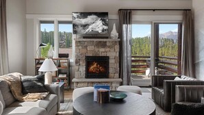 A cozy living room with a stone fireplace, a comfortable sofa, a round coffee table, and a view of a mountainous landscape through large windows. - Saddle Ridge B5 3 Bedroom Townhouse (Big Sky)