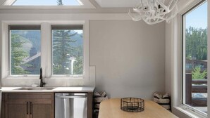 Una cocina moderna con lavavajillas, una mesa redonda y dos ventanas que ofrecen vistas a árboles y una montaña.