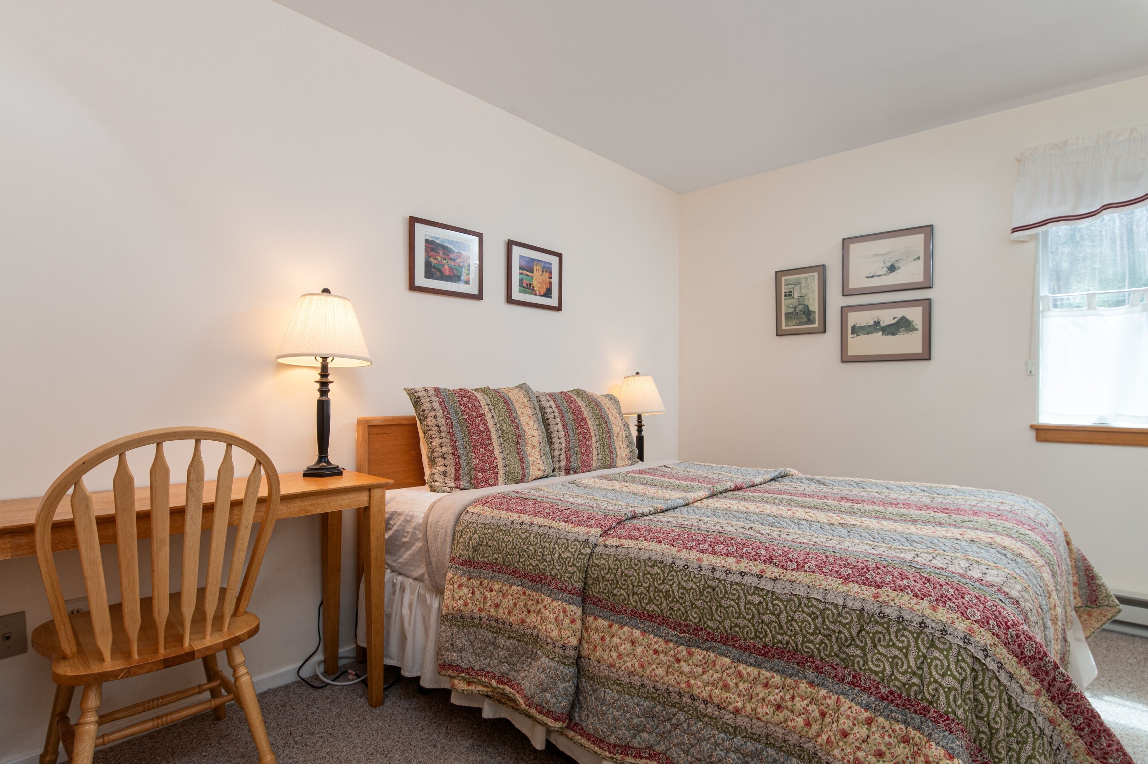 A bedroom with a bed, a wooden chair, two bedside lamps, and three framed pictures on the wall.