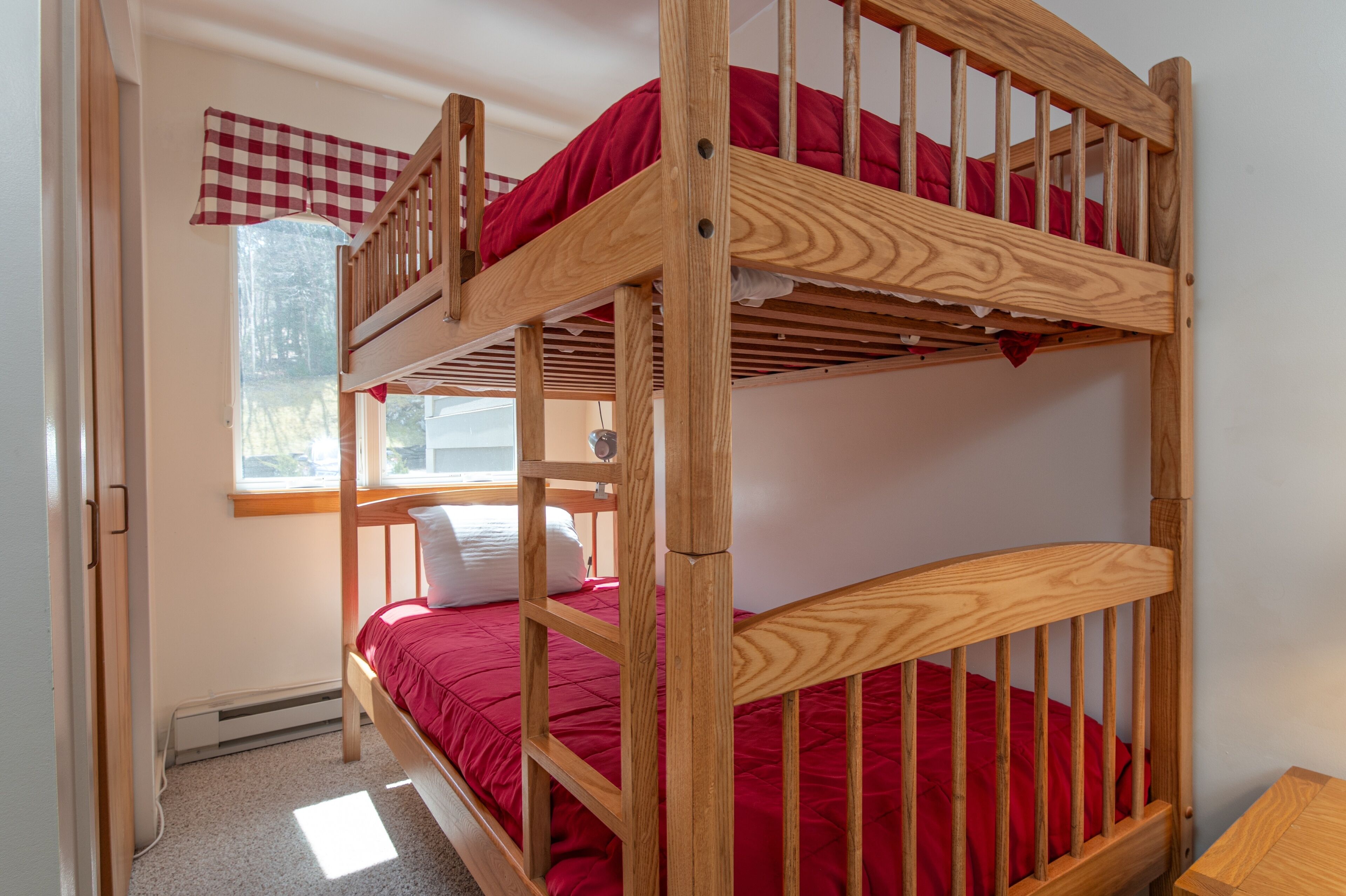 A bunk bed with a red mattress, a window with a checkered curtain, and a wooden table beside the bed.