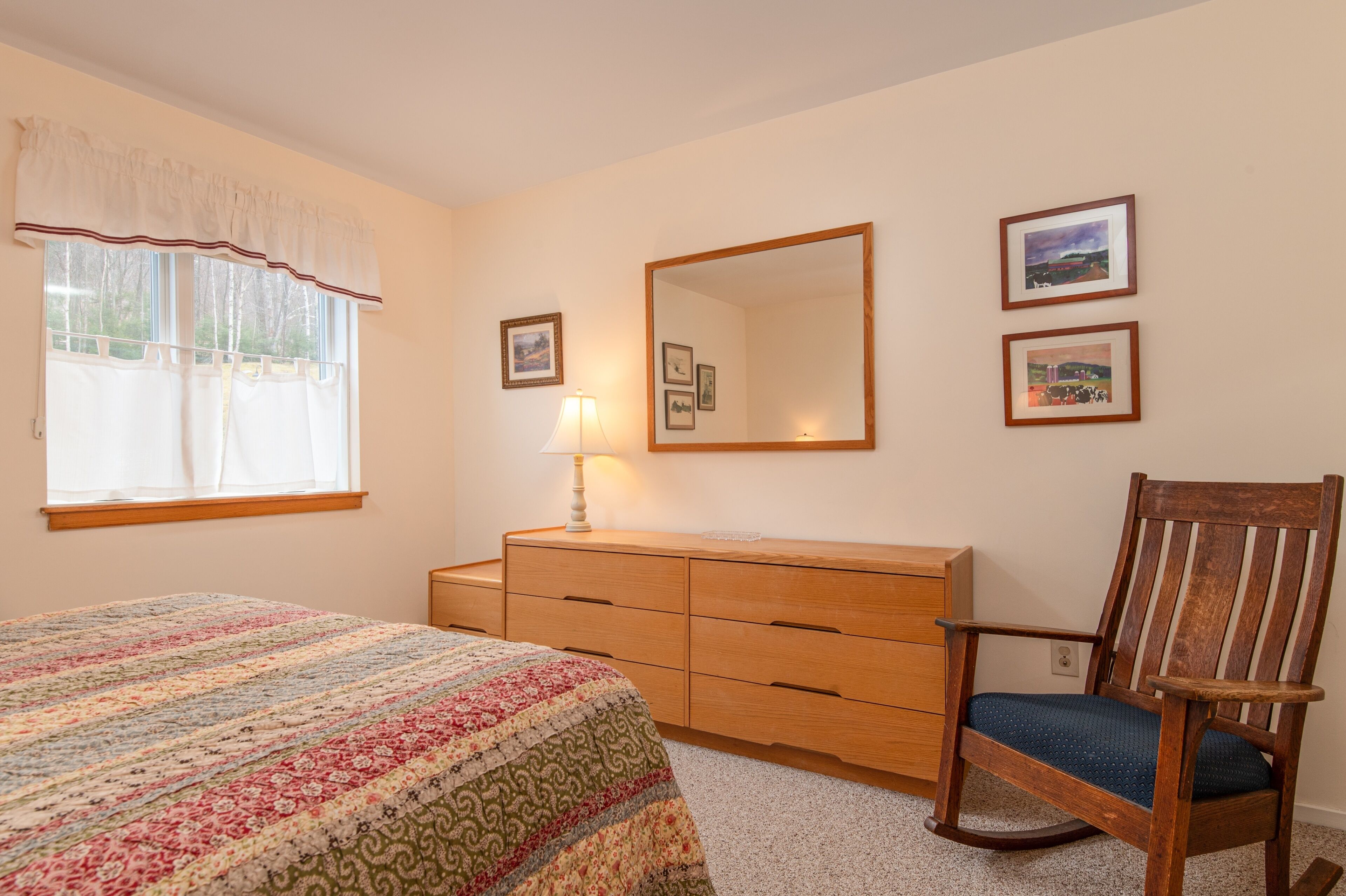 A bedroom with a bed, a wooden dresser, a rocking chair, a window with curtains, and wall-mounted pictures.