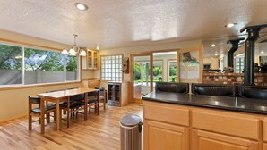 A kitchen with wooden cabinets, a stainless steel trash can, and a dining area with a table and chairs. - Otter Run 3 Bedroom Home (Bend)