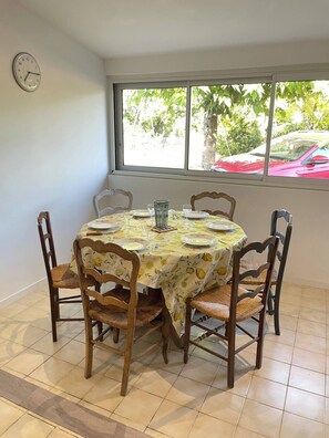 Zona pranzo con tavolo rotondo apparecchiato per quattro persone, circondato da sedie in legno, un orologio a parete e una finestra con vista su alberi e un'auto rossa all'esterno.