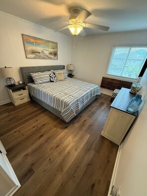 A bedroom with a bed, a ceiling fan, a nightstand, a lamp, a window, and a wooden floor. - Cozy Unit at Oceanfront Complex, Pool & Steps from Ocean by Williamson Realty (Ocean Isle Beach)
