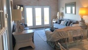 A cozy room with a wooden ceiling, a large window with a view, a white sofa, a small table with a lamp, and a decorative wall piece.