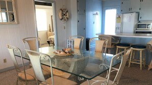 A dining area with a glass table surrounded by white metal chairs, a decorative wall hanging, and a kitchen area with cabinets and appliances.
