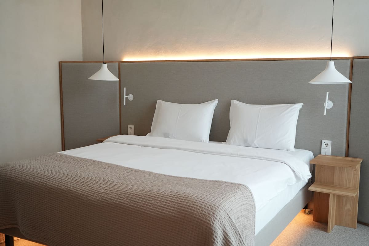 Una cama bien hecha con sábanas blancas y una manta marrón, una mesita de noche de madera con cajón, y dos lámparas colgantes sobre la cama.