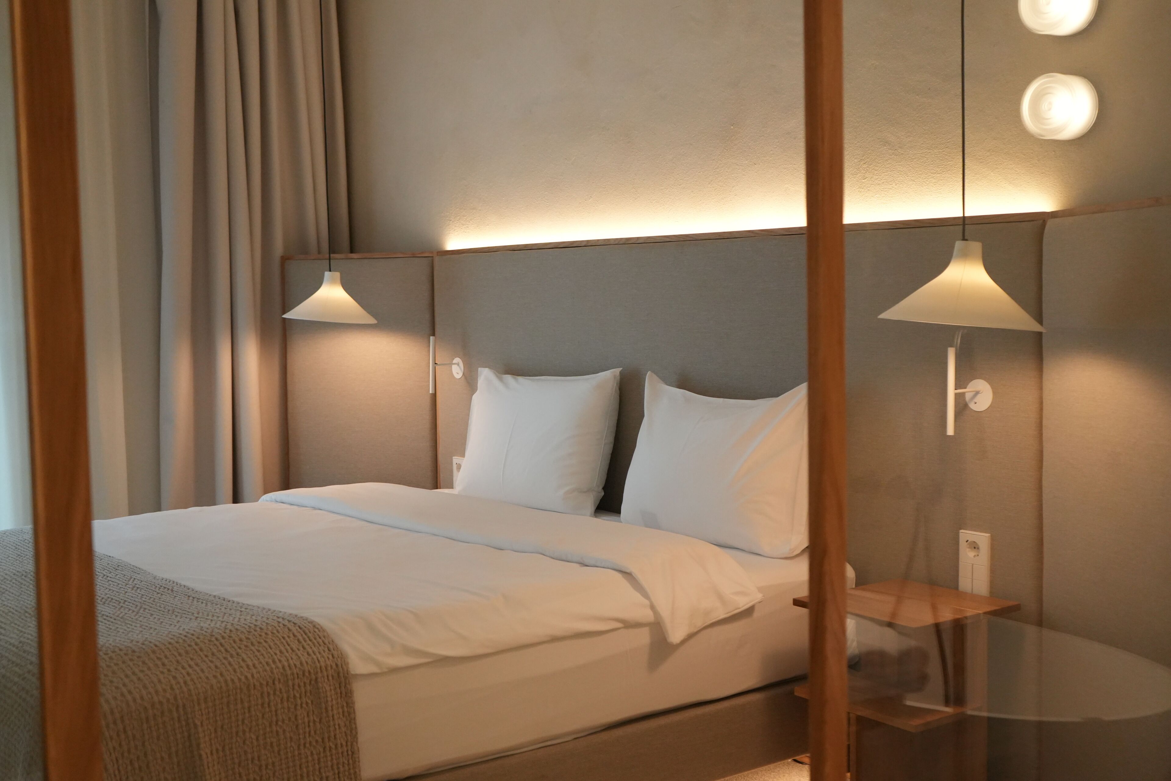 Una cama bien hecha con sábanas blancas, cabecera con iluminación integrada y una mesita de noche con lámpara.
