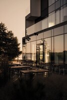 Un espace extérieur aménagé avec des tables et des chaises, de grandes fenêtres qui reflètent le paysage environnant et une vue sur une étendue d’eau.