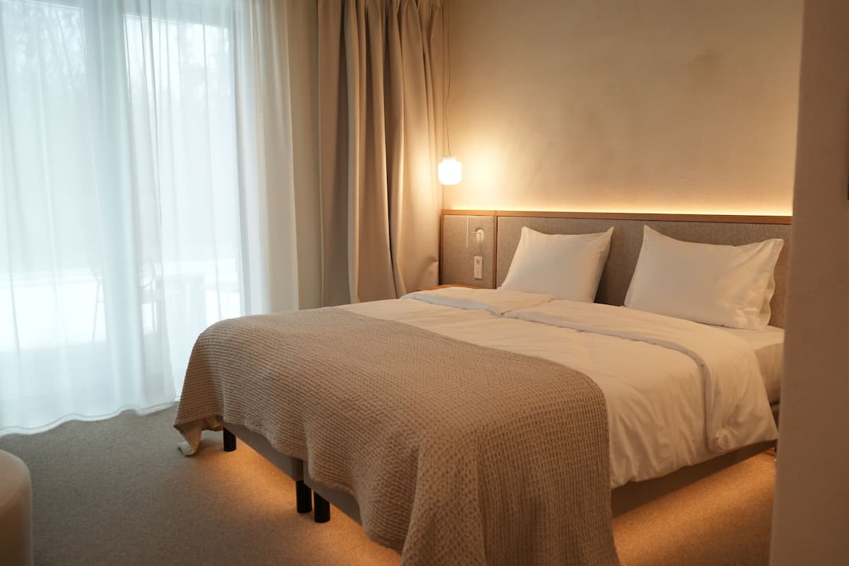 Una cama bien hecha con cabecera, lámpara de noche y ventana con cortinas traslúcidas.