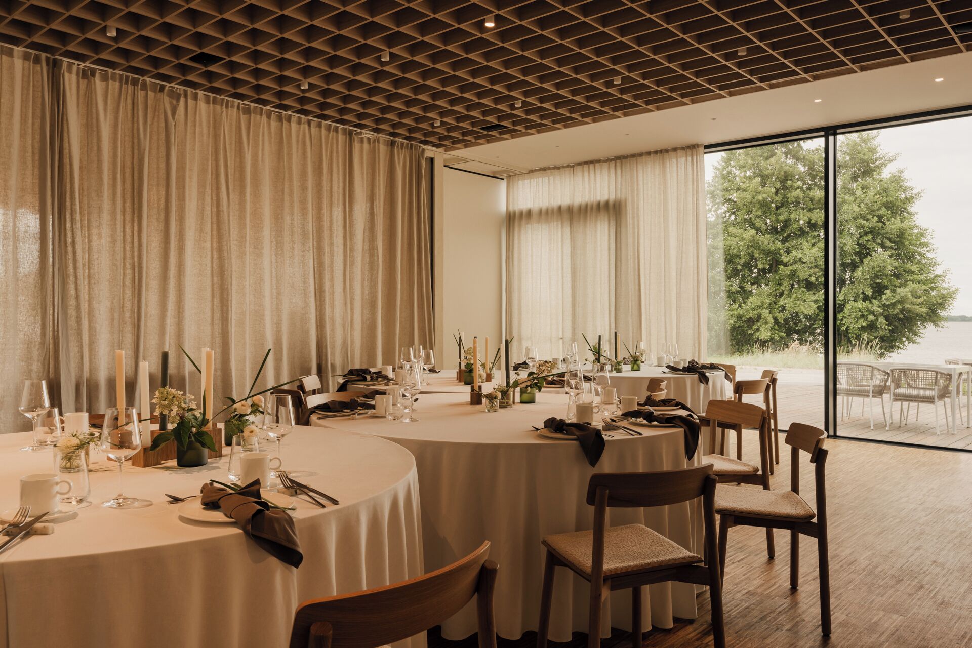 A dining area with round tables set for a meal, large windows offering a view of the outdoors, and a ceiling with a grid pattern.