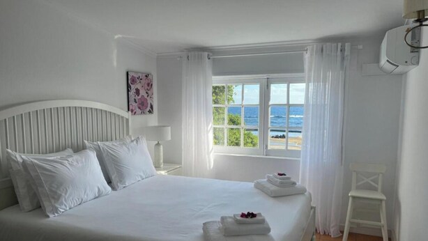 Quarto branco com cama, mesa de cabeceira, janela com vista para o mar e uma cadeira branca.
