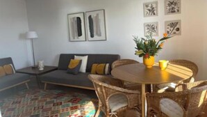 Uma sala de estar com sofá, mesa de centro com um vaso, e uma mesa de jantar com cadeiras. Há obras de arte nas paredes acima do sofá e da mesa de jantar.