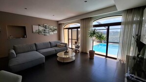 A modern living room with a grey sofa, a round coffee table, and a large window overlooking a pool. - Aurelia Gozo BnB (Xaghra)