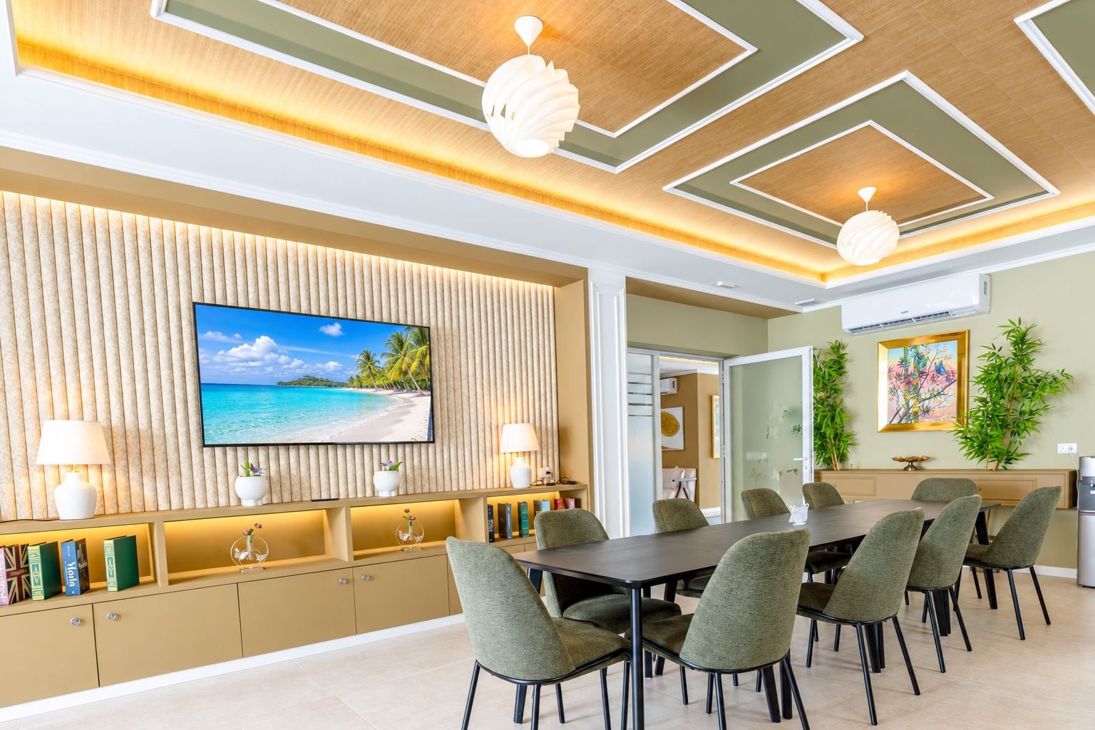 Ein moderner Essbereich mit großem Tisch, mehreren Stühlen, einem an einer Holzvertäfelung montierten Fernseher und Blick auf einen Strand durch das Fenster.