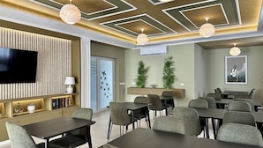 A modern hotel room with a flat-screen TV, a sofa, a dining area with tables and chairs, and a ceiling with geometric patterns. - Casual Green Village Bayahibe by Casual Hoteles (Bayahíbe)